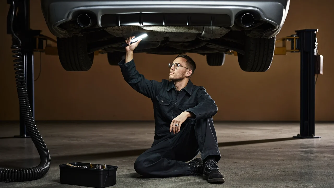 Illustration: Mechaniker prüft die Fahrzeugunterseite mit Handlampe auf der Hebebühne