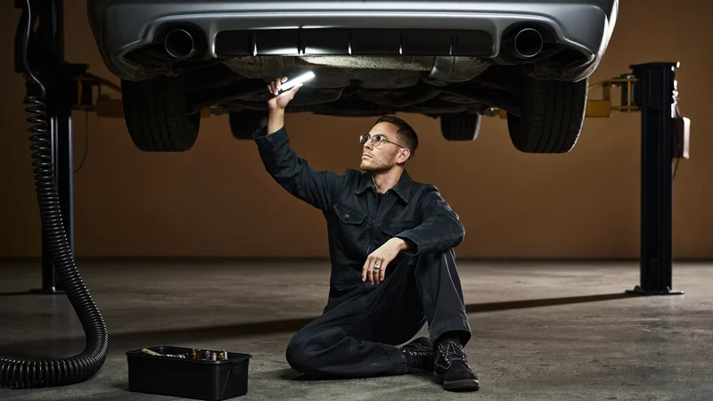 Illustration: Mechaniker prüft die Fahrzeugunterseite mit Handlampe auf der Hebebühne